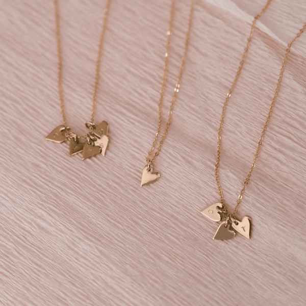 Three delicate gold pendant chain necklaces each with small flat heart charms. Some of the hearts are engraved with single initials, and all of the necklaces are laying on a textured light beige surface.