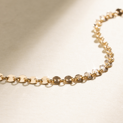 A close-up of a gold bracelet consisting of multiple round, flat discs connecting, set against a neutral background with natural shadows.