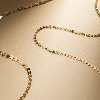 A gold chain necklace made up of flat, circular discs linked together, set against a textured neutral background.