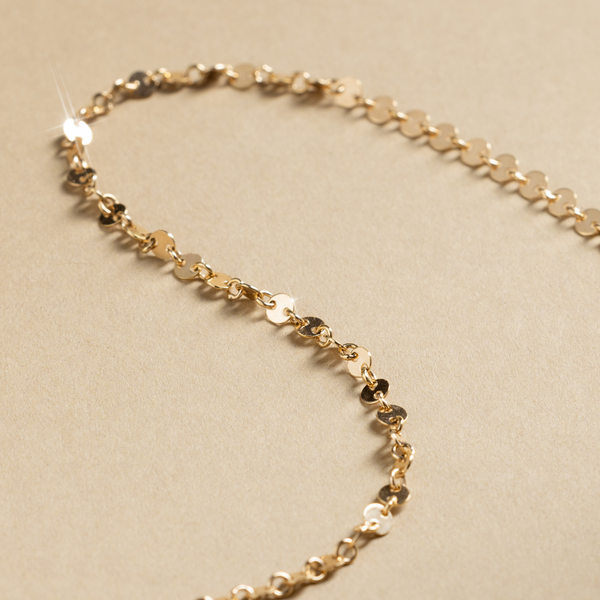 A gold necklace made up of flat, circular discs linked together, set against a textured beige background.