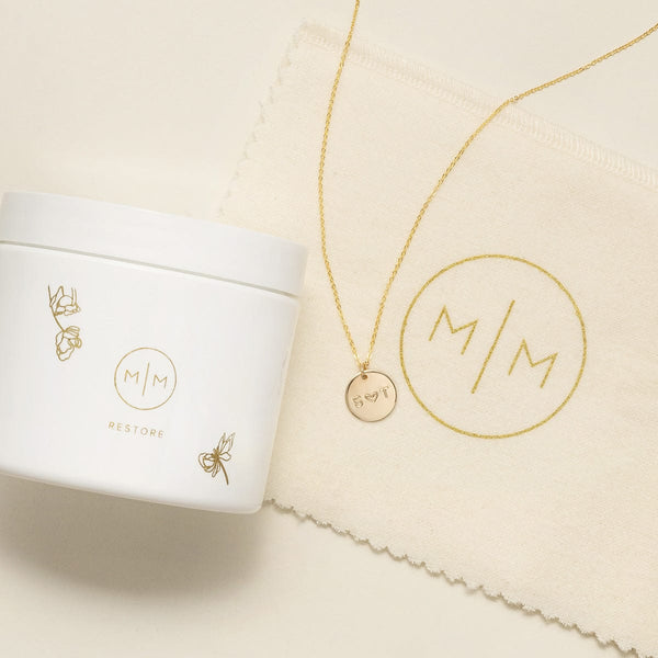 A gold necklace with a round engraved pendant displayed next to a white jar of jewelry cleaner and a cream microfiber cloth, all branded with the monogram M/M. The necklace is laid on a light beige surface.