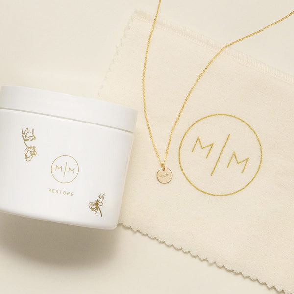 A gold necklace with a round engraved pendant displayed next to a white jar of jewelry cleaner and a cream microfiber cloth, all branded with the monogram M/M. The necklace is laid on a light beige surface.