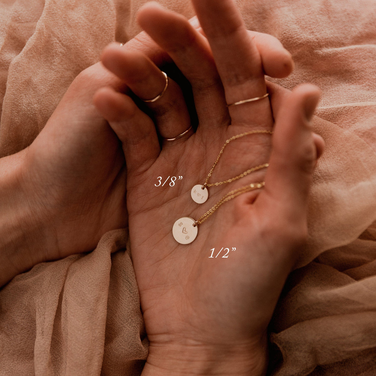 Two hands resting on a light pink linen cloth, holding two delicate gold pendant chain necklaces with engraved discs in different sizes. Text on the screen shows the measurements of the discs: '3/8"' and '1/2"'.