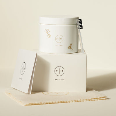 A white box with a white jar and small cleaning brush on top, each with an M|M logo and the word 'RESTORE'. They are both sitting on a cream polishing cloth and shown against a solid light beige background.