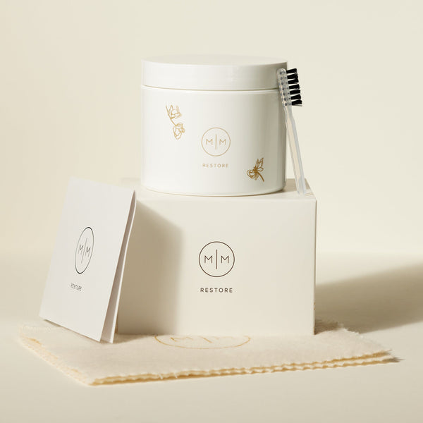 A white box with a white jar and small cleaning brush on top, each with an M|M logo and the word 'RESTORE'. They are both sitting on a cream polishing cloth and shown against a solid light beige background.