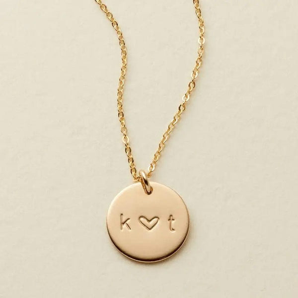 A delicate gold pendant chain necklace with an engraved disc reading 'k ♥ t'.
