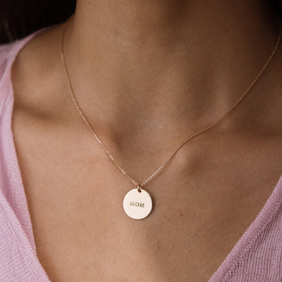 A close-up of a person's neck and chest wearing a delicate gold chain necklace with a round, engraved disc pendant that reads 'MOM'. The person is wearing a light pink knit top.