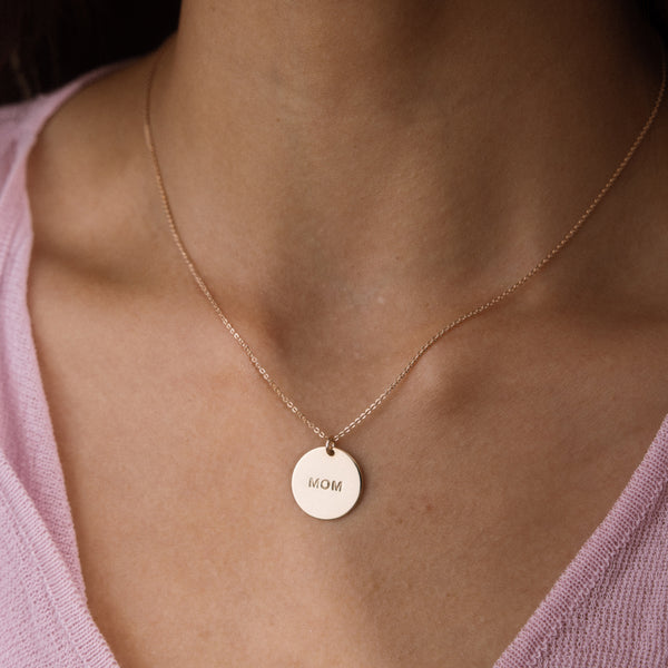 A close-up of a person's neck and chest wearing a delicate gold chain necklace with a round, engraved disc pendant that reads 'MOM'. The person is wearing a light pink knit top.