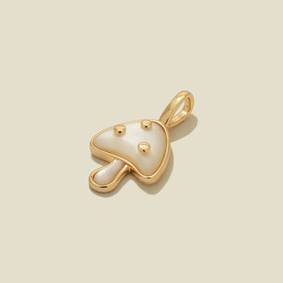 A small mushroom-shaped pendant charm with a gold-tone metal outline and a glossy white stone inlay, featuring three small gold dots on the cap, centered on a light beige background.