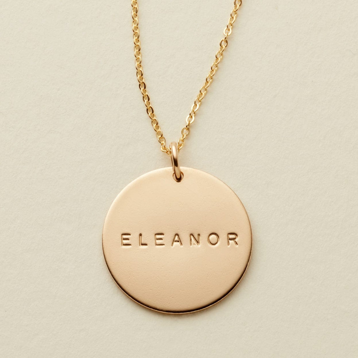 A gold chain necklace with a round gold disc pendant engraved with the name 'ELEANOR', displayed on a textured light beige background.