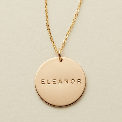 A gold chain necklace with a round gold disc pendant engraved with the name 'ELEANOR', displayed on a textured light beige background.