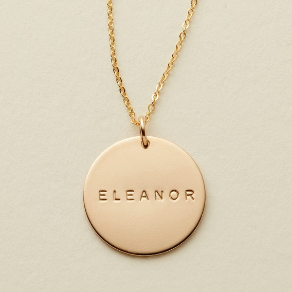 A gold chain necklace with a round gold disc pendant engraved with the name 'ELEANOR', displayed on a textured light beige background.