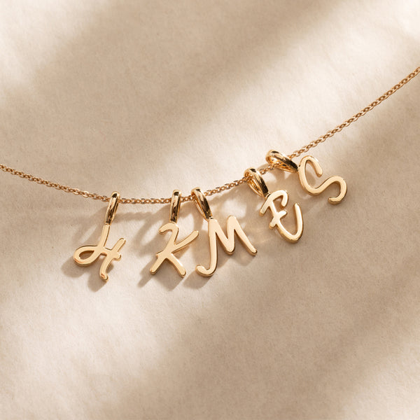 Close-up of a gold chain necklace featuring multiple individual cursive initial charms spaced along the chain against a neutral background.