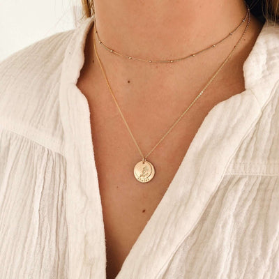 A close-up of a person in a white linen top wearing a dainty gold choker with stationary round beads and a longer pendant chain necklace featuring two stacked round flat discs. The top disc shows an engraved flower, and the larger bottom has the word 'violet' etched along the bottom.