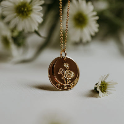 A close-up of a delicate gold pendant chain with a single charm, featuring two round flat discs stacked together. The smaller disc on top is engraved with a floral design, and the larger disc in the back is stamped with the letters 'M.B.M.' along the bottom.