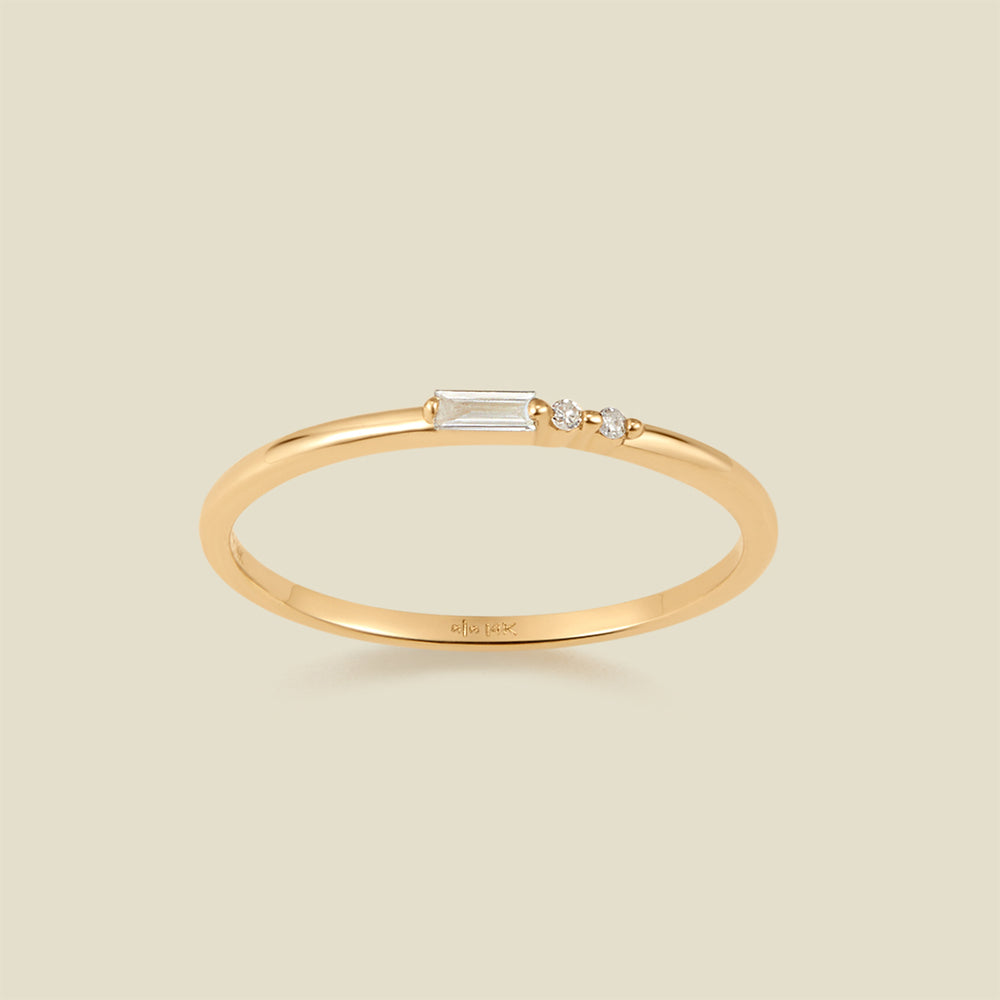A delicate yellow gold ring featuring a baguette-cut clear diamond set next to two small clear round-cut diamonds, set against a solid light beige background.