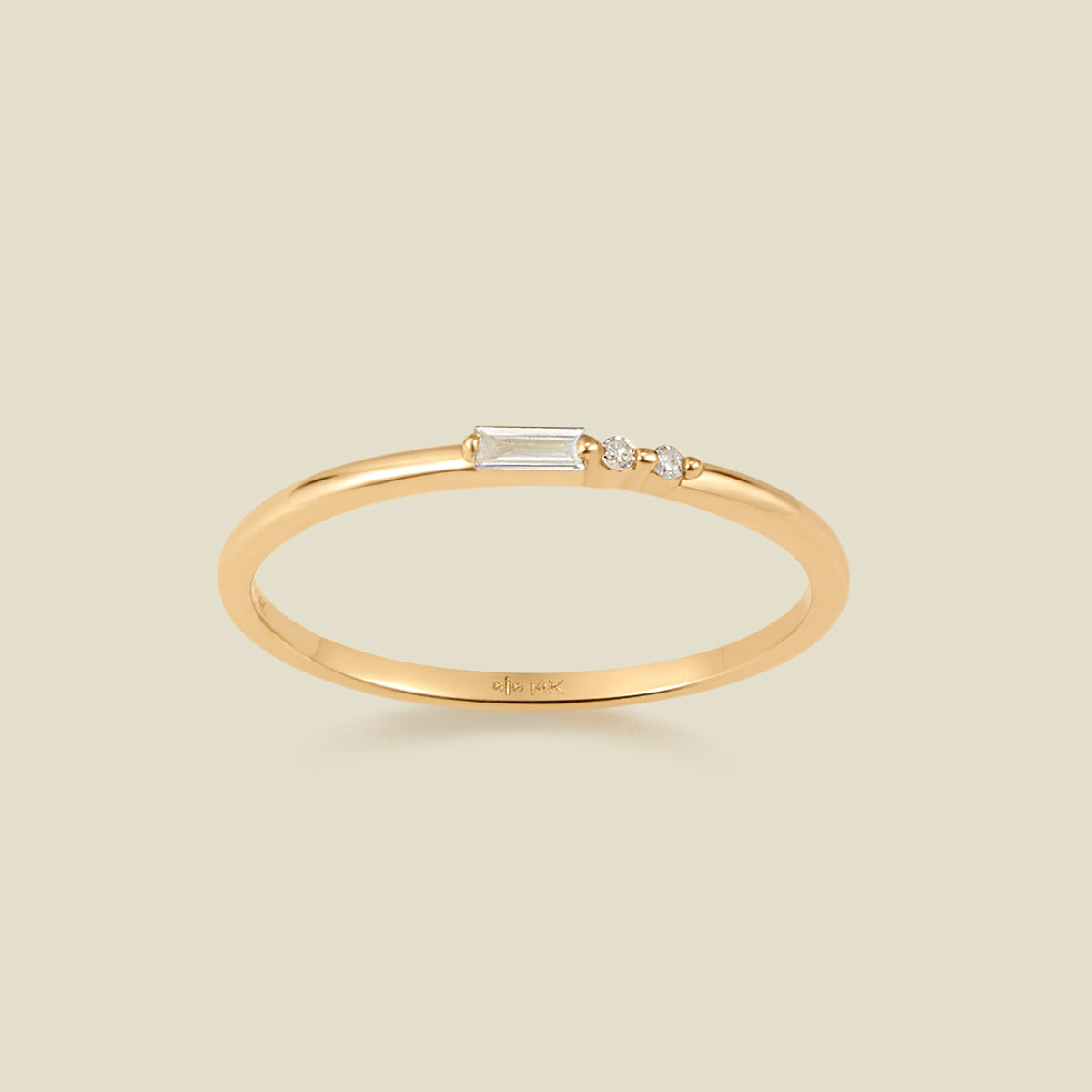 A delicate yellow gold ring featuring a baguette-cut clear diamond set next to two small clear round-cut diamonds, set against a solid light beige background.