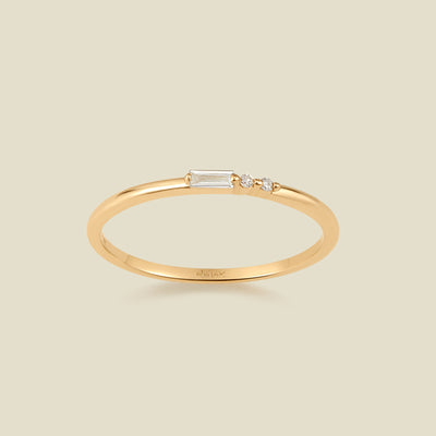 A delicate yellow gold ring featuring a baguette-cut clear diamond set next to two small clear round-cut diamonds, set against a solid light beige background.