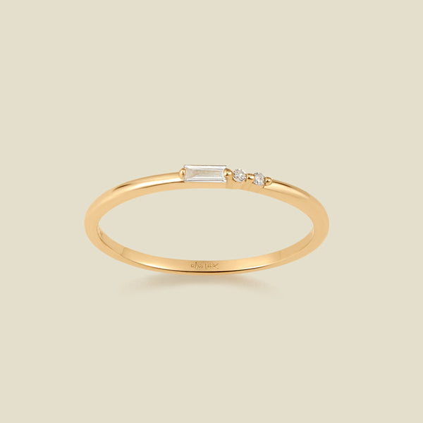 A delicate yellow gold ring featuring a baguette-cut clear diamond set next to two small clear round-cut diamonds, set against a solid light beige background.