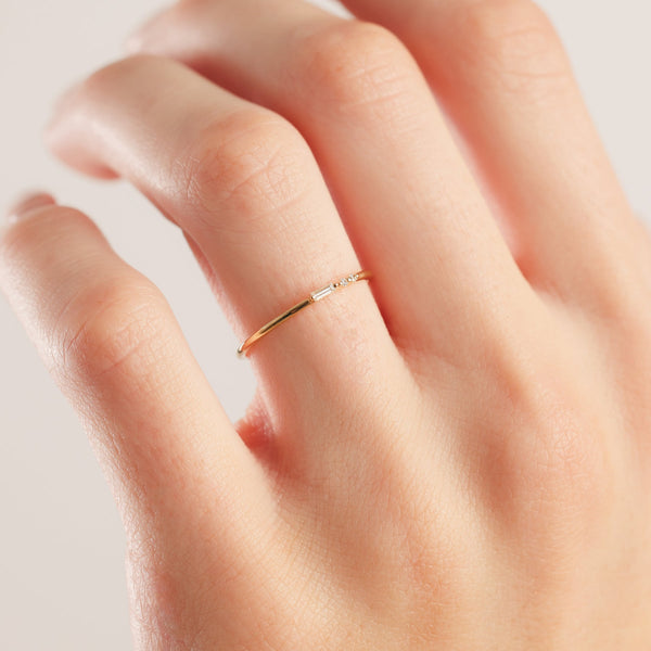 A close-up of a person's hand showcasing a thin gold band ring with an east-west clear baguette diamond followed by two small clear round-cut diamonds, shown on a white background.