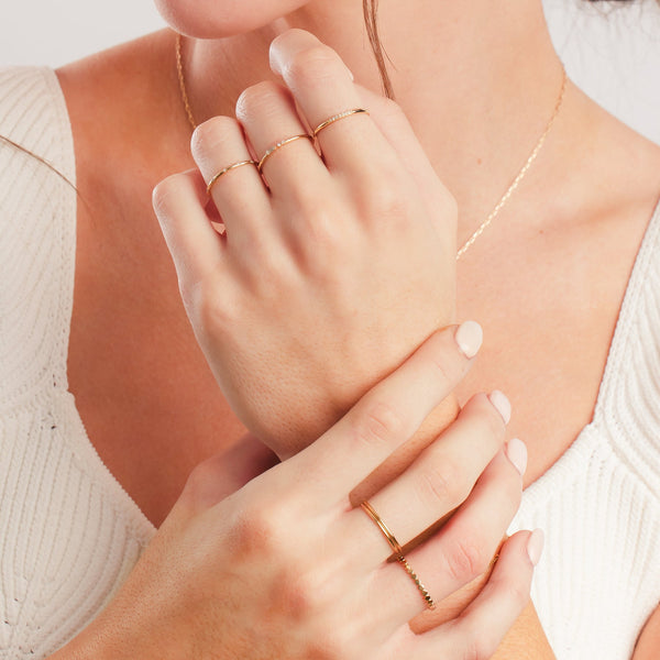 A close-up of a person's hands held up in front of their chest, showing multiple gold rings in different styles, some with clear gemstone accents, paired with a white knit top and shown on a solid white background.