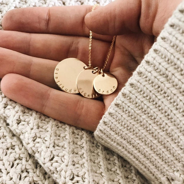 A person wearing a light beige knit sweater holding a gold disc pendant necklace with three different sized round charms on a chain, each engraved with a word: gratitude, dignity, and grace, shown on a light neutral background.