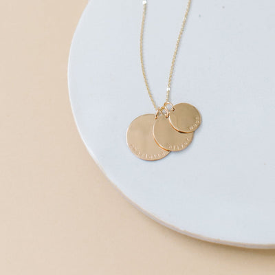 A gold disc pendant necklace with three  different sized round charms on a chain, each engraved with a personalized name or word: charlotte, oliver, and west, shown on a bright white circular surface with a solid light beige background.