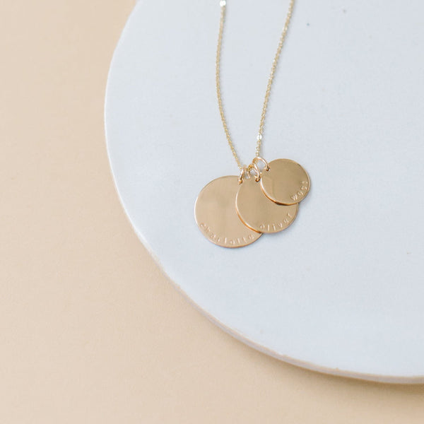A gold disc pendant necklace with three  different sized round charms on a chain, each engraved with a personalized name or word: charlotte, oliver, and west, shown on a bright white circular surface with a solid light beige background.