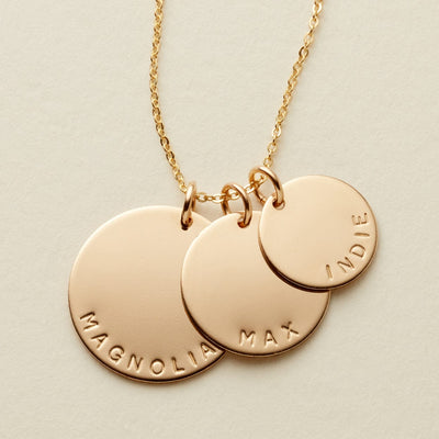 A gold disc pendant necklace with three  different sized round charms on a chain, each engraved with a personalized name or word: MAGNOLIA, MAX, and INDIE, shown on a light neutral background.