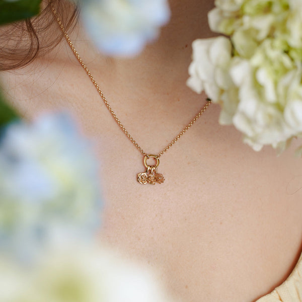 A woman wearing a gold charm necklace with a round ring in the center, holding three small flower charms, with white and blue flowers blurred in the foreground.