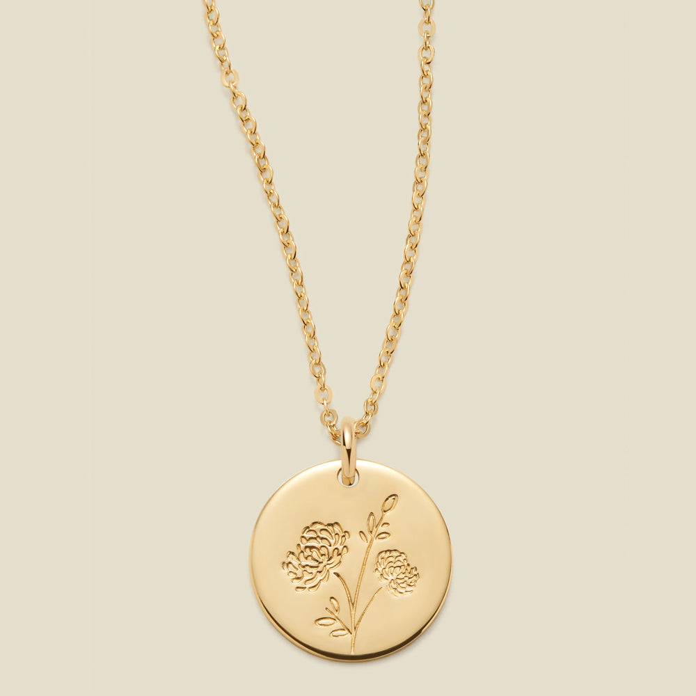 A gold chain necklace with a round gold disc pendant engraved with a chrysanthemum floral design, shown against a solid light beige background.