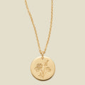 A gold chain necklace with a round gold disc pendant engraved with a chrysanthemum floral design, shown against a solid light beige background.