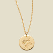 A gold chain necklace with a round gold disc pendant engraved with a chrysanthemum floral design, shown against a solid light beige background.