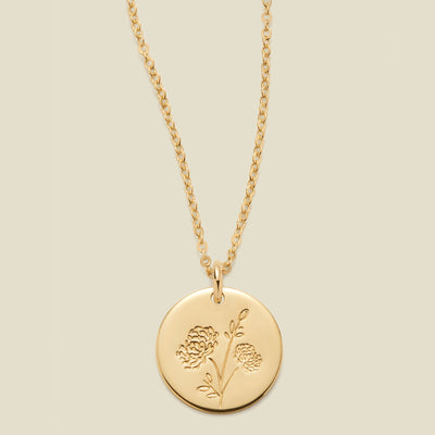 A gold chain necklace with a round gold disc pendant engraved with a chrysanthemum floral design, shown against a solid light beige background.