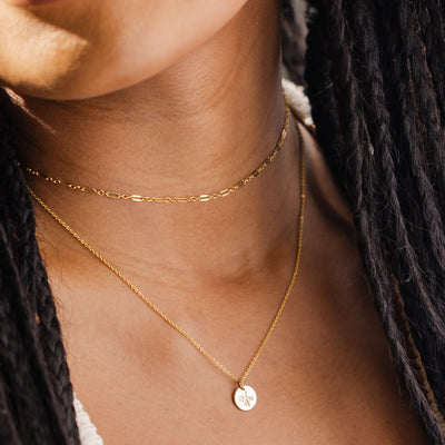 A close-up of a woman wearing a thin textured gold choker layered with a delicate gold pendant chain featuring a round flat disc with a floral engraving.