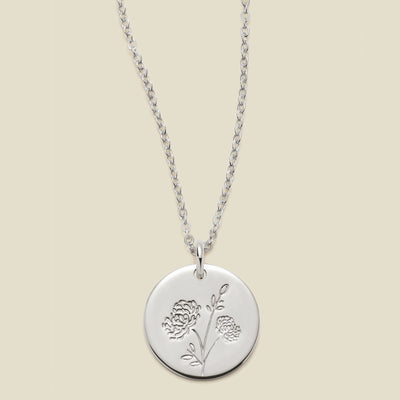 A silver chain necklace with a round silver disc pendant engraved with a chrysanthemum floral design, shown against a solid light beige background.