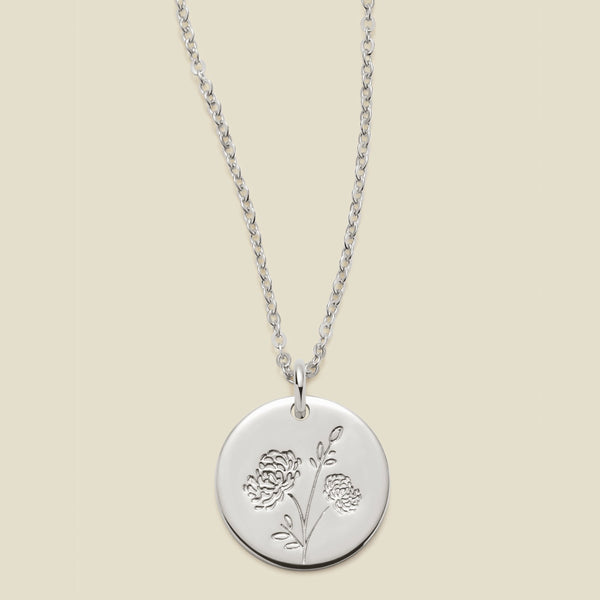 A silver chain necklace with a round silver disc pendant engraved with a chrysanthemum floral design, shown against a solid light beige background.