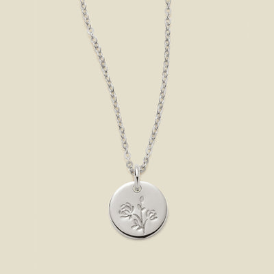A silver chain necklace with a round silver disc pendant engraved with a chrysanthemum floral design, shown against a solid light beige background.