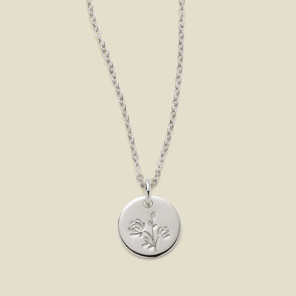 A silver chain necklace with a round silver disc pendant engraved with a chrysanthemum floral design, shown against a solid light beige background.
