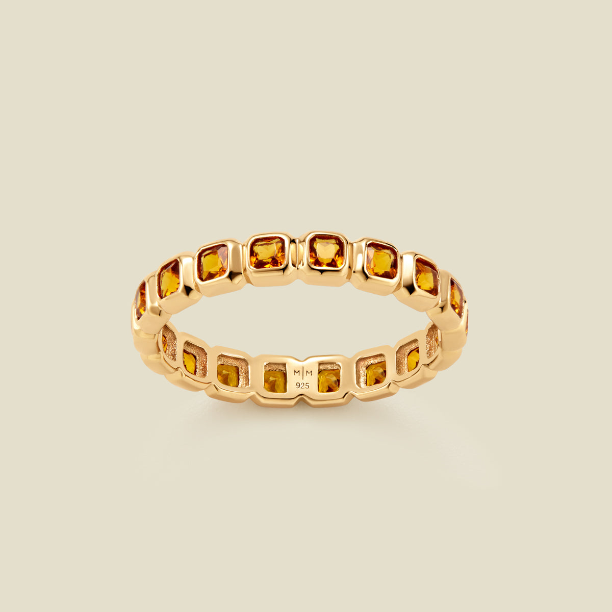 A gold eternity ring featuring a continuous row of square cut amber gemstones, shown on a solid light beige background.