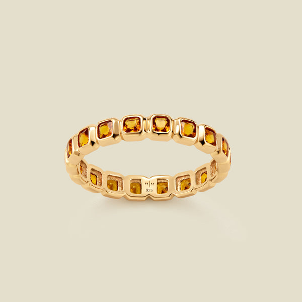 A gold eternity ring featuring a continuous row of square cut amber gemstones, shown on a solid light beige background.