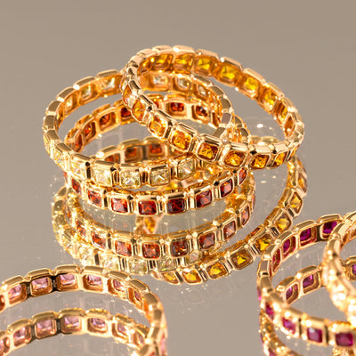A stack of three gold eternity band rings, each with either amber, clear, or red square cut gemstones in a single continuous row. Their are other rings blurred in the foreground, and the stack is sitting on a mirror surface.