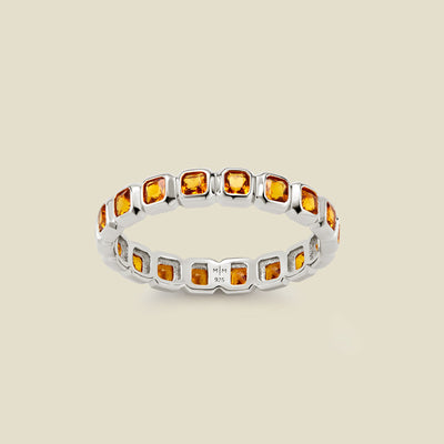 A silver eternity ring featuring a continuous row of square cut amber gemstones, shown on a solid light beige background.