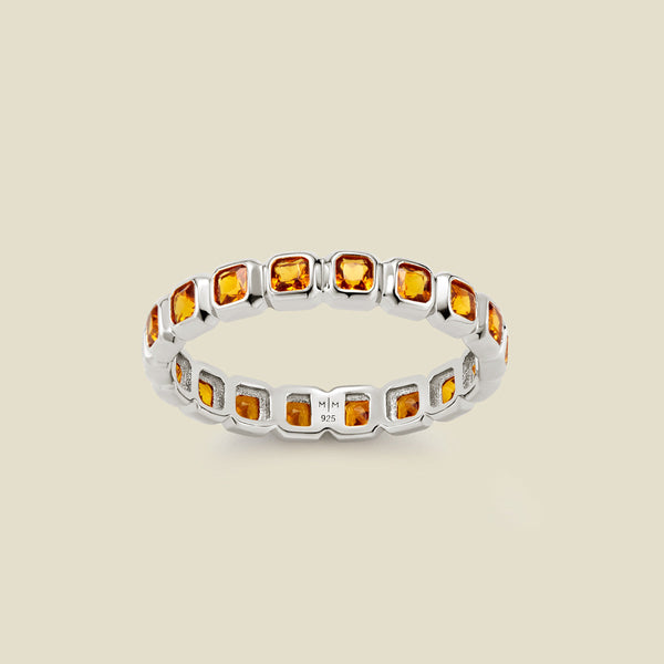 A silver eternity ring featuring a continuous row of square cut amber gemstones, shown on a solid light beige background.