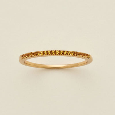 A gold ring featuring a single row of small round-cut amber gemstones, photographed on a clean, solid, warm off-white background.