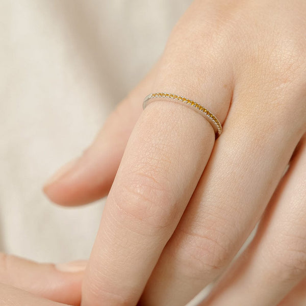 A close-up of a hand wearing a thin silver band ring with a single row of small amber gemstones across the front, set against a light beige background.