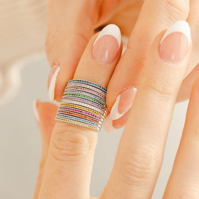A close-up of a finger wearing 12 stacked thin silver rings, each with a single row of small gemstones in varying colors.