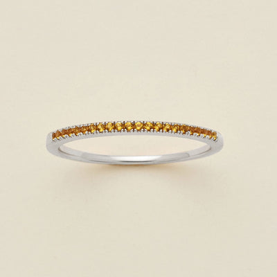 A silver ring featuring a single row of small round-cut amber gemstones, photographed on a clean, solid, warm off-white background.
