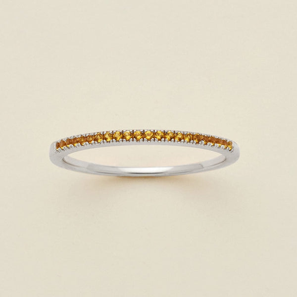 A silver ring featuring a single row of small round-cut amber gemstones, photographed on a clean, solid, warm off-white background.