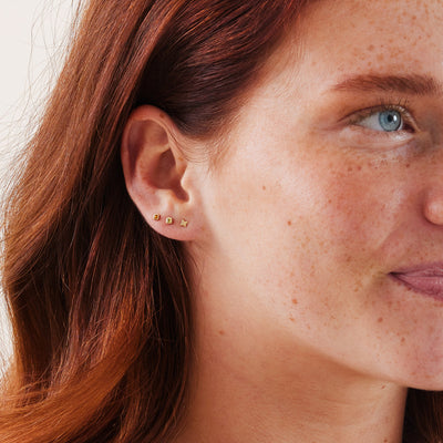 A close-up of a person's ear wearing a gold star stud earring, and two gold square bezel-set gemstone studs, one with an amber stone and one with a light green stone.
